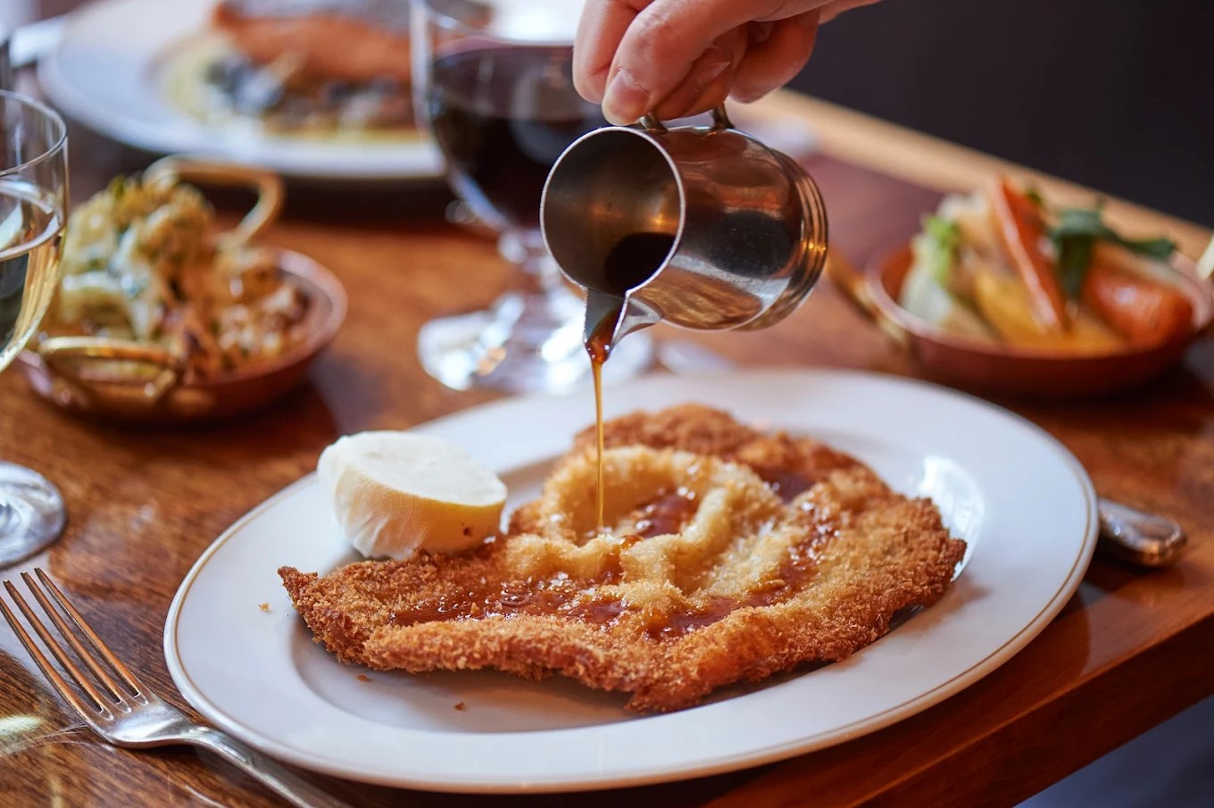 A schintzel being covered in gravy sits in an elegant ceramic plate surrounded by other platters on a dining table.