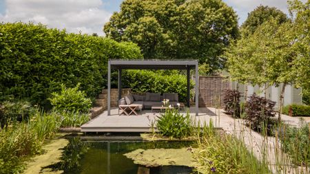 a pergola in a backyard with a natural pool/pond with an L-shaped sofa, a large hedge on one side and a garden path on the other side flanked by trees