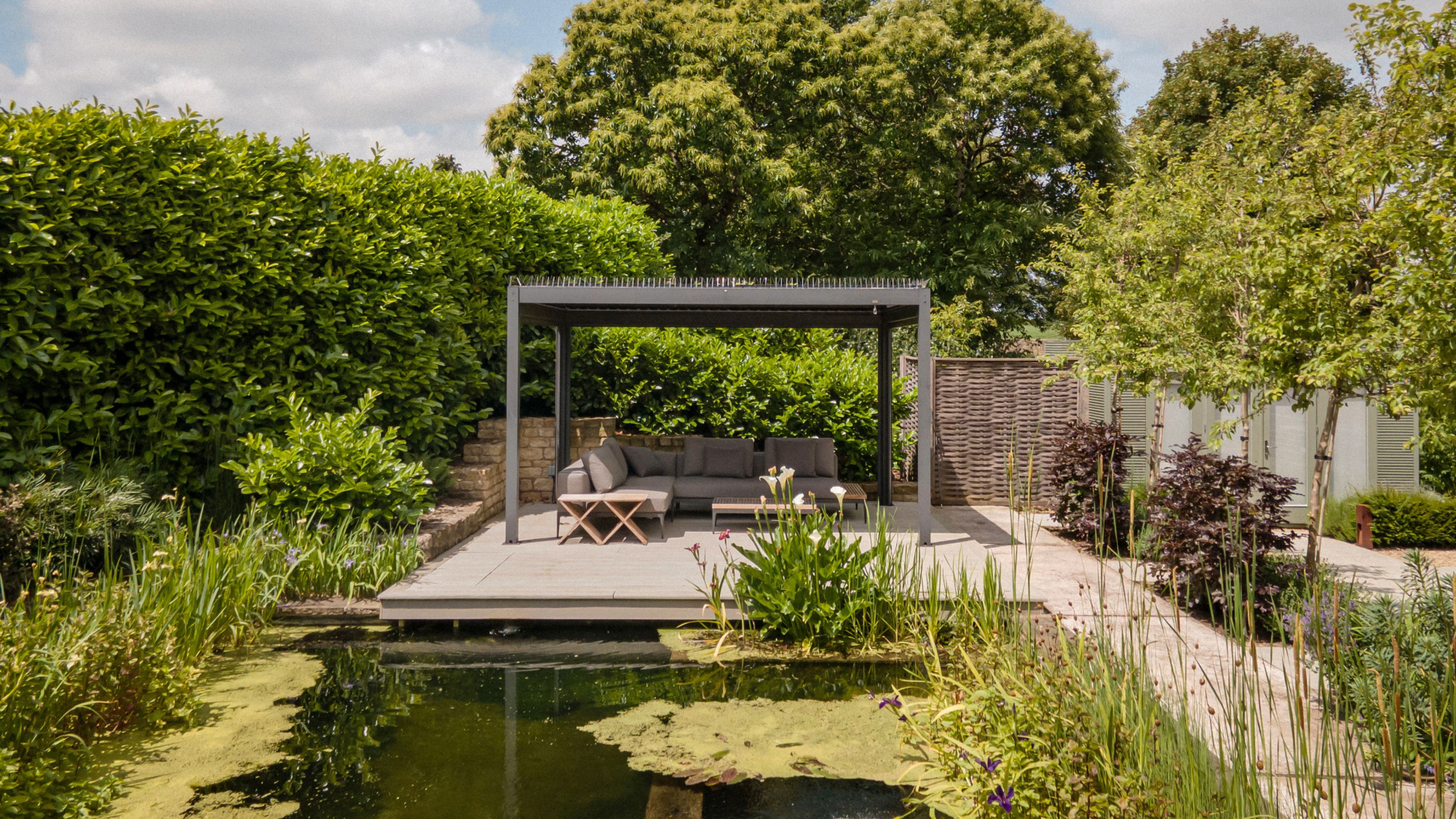 a pergola in a backyard with a natural pool/pond with an L-shaped sofa, a large hedge on one side and a garden path on the other side flanked by trees