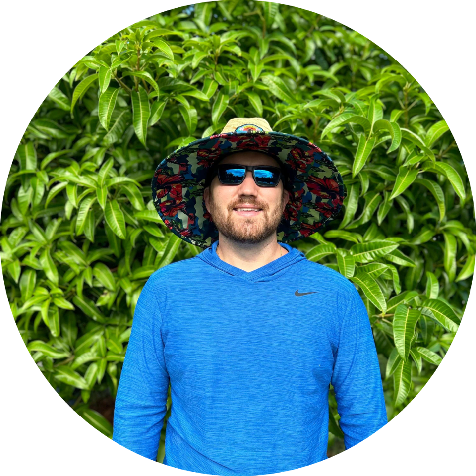 Head shot of male in blue top, hat and sunglasses, stood in front of lush greenery