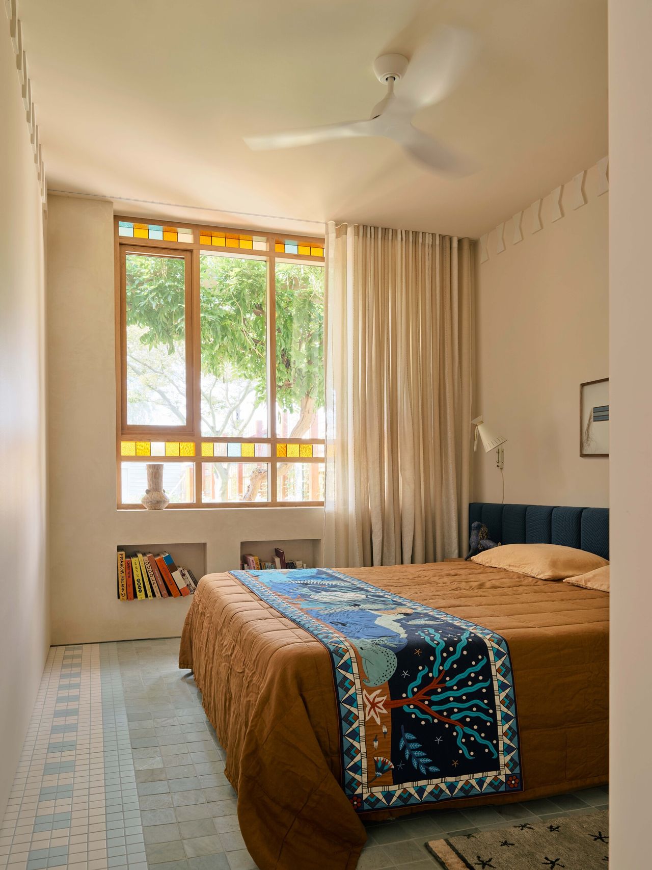 modern bedroom with hempcrete walls and modern wall molding, extended upholstered headboard with orange bedcover and colorful blanket, floating timber side table with statue, wall light, artwork above bed, and timber framed windows with stained glass detail and alcove niches built into wall and tiled floor