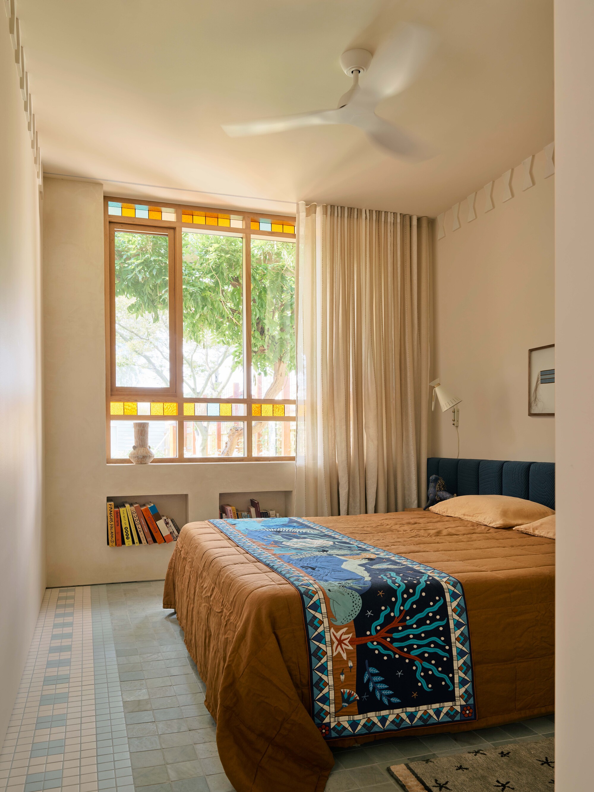 modern bedroom with hempcrete walls and modern wall molding, extended upholstered headboard with orange bedcover and colorful blanket, floating timber side table with statue, wall light, artwork above bed, and timber framed windows with stained glass detail and alcove niches built into wall and tiled floor