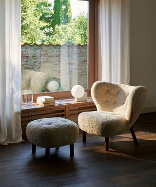 Sheepskin lounge chair and matching footstool beside window, low sideboard decorated with lamp, books and ornaments, dark wooden floor, sheer curtains