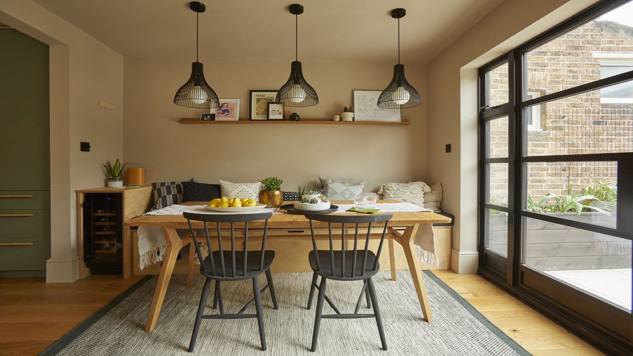 small dining room area with bench seating, two black chairs, open shelving, three wire pendant lights, rug, crittall doors to garden, cushions, fruit on table, table runner, glasses, artwork, kitchen to the left