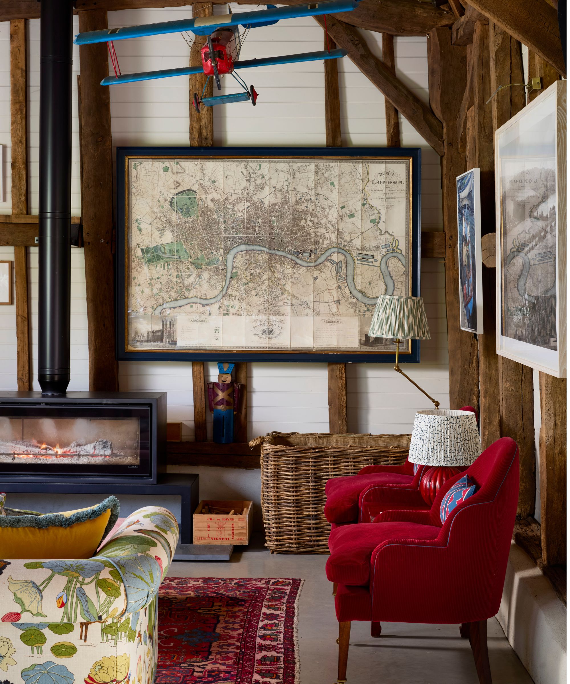 a rustic beamed barn with red armchairs, a fireplace, a large framed map and a red and blue model plane suspended from the ceiling