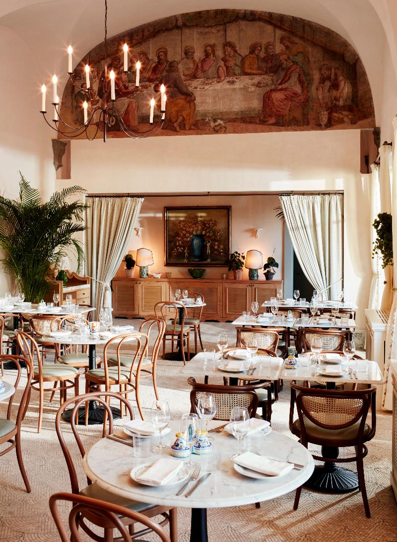 A naturally lit dining room with bistro-style marble and wrought iron tables and wooden chairs, pale green curtains, trailing plants, warm lighting, and white-washed walls with a red-tinted figurative fresco.