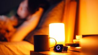 A warm amber night light on bedside table next to apple watch and mug. Person laying in bed in blurred background.