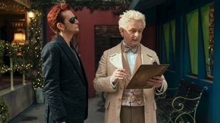 David Tennant in a black suit and sunglasses as Crowley stands alongside Michael Sheen in a cream suit with a clipboard as Aziraphale in Good Omens season 2.