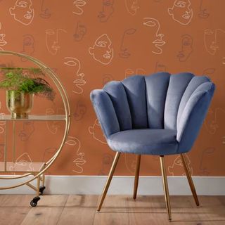 Terracotta wallpaper featuring faces on a wall with a blue velvet chair in front of it