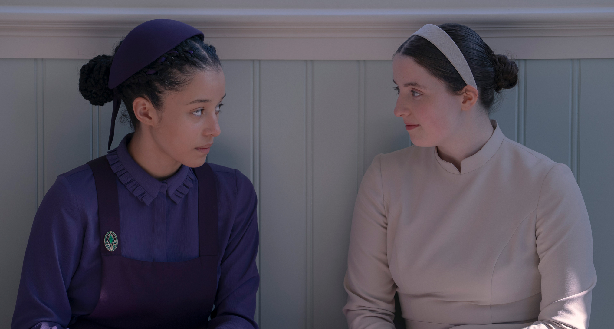 chase infiniti as agnes wearing a purple dress and lucy halliday as daisy wearing a white dress as they tlak to each other sitting next to a blue wall in the testaments