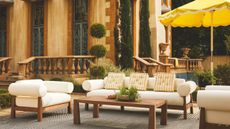 an outdoor seating area with a white accent chair, a white couch with yellow pillows, a wooden coffee table, a striped rug, and a yellow umbrella