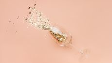 A tipped over champagne glass with gold glitter pouring out on a pink background