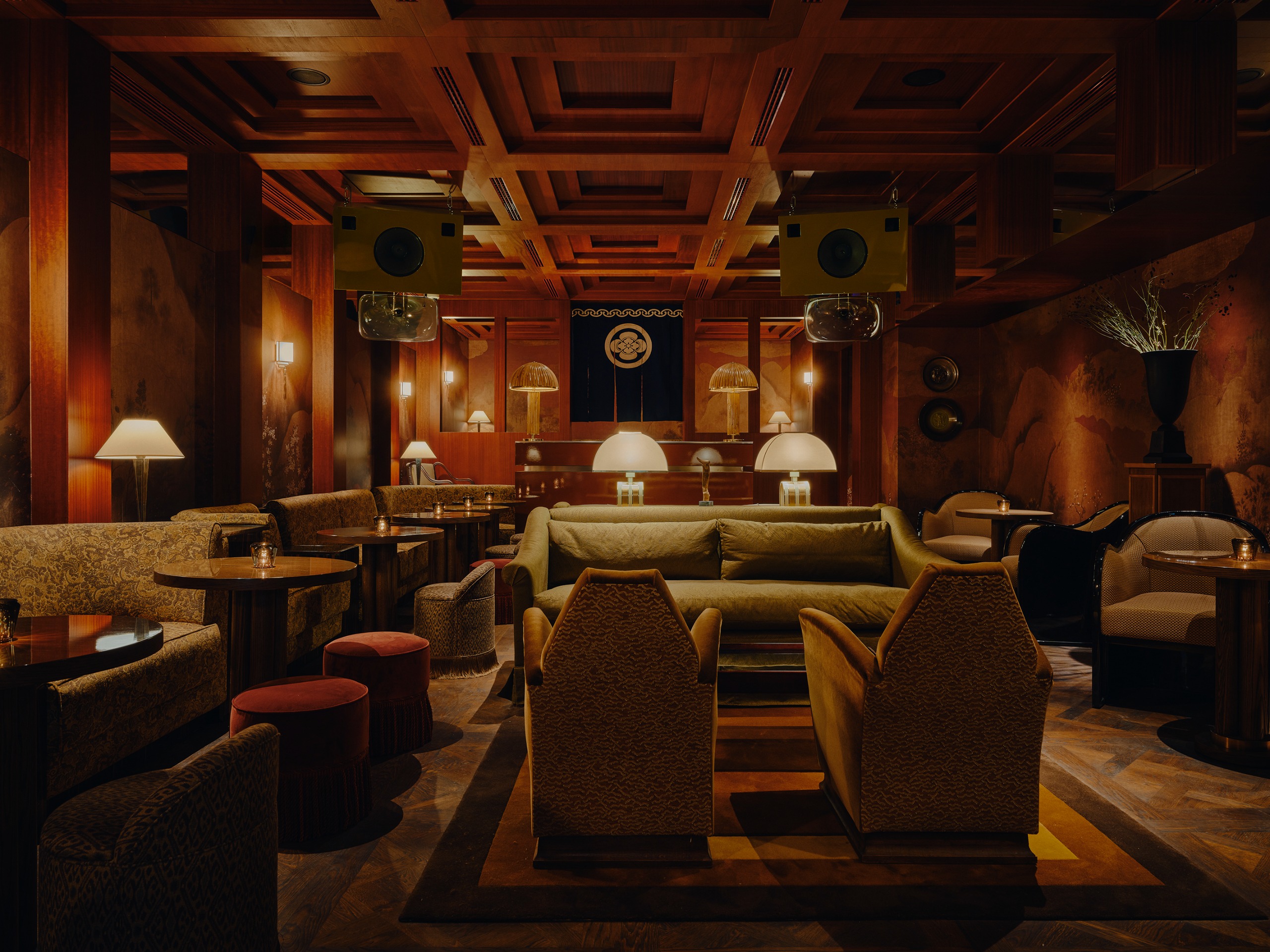 A speakeasy-style bar with low-lighting, a rich, moody color palette with sofas, armchairs and pouffes and two large green speakers suspended from the red-painted layered ceiling
