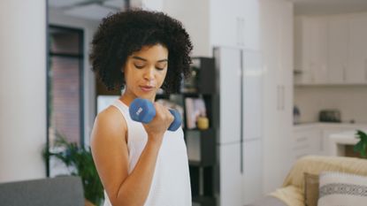 woman curling a blue dumbbell to her biceps, wearing a white vest in a home setting