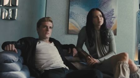 From left to right: Josh Hutcherson as Peeta sitting with his arms resting against the back of a couch and Jennifer Lawrence as Katniss sitting next to him. 