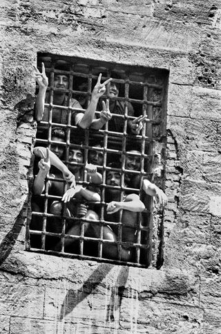 Black and white photograph of a fenced window, with several arms outstretched through, making peace signs