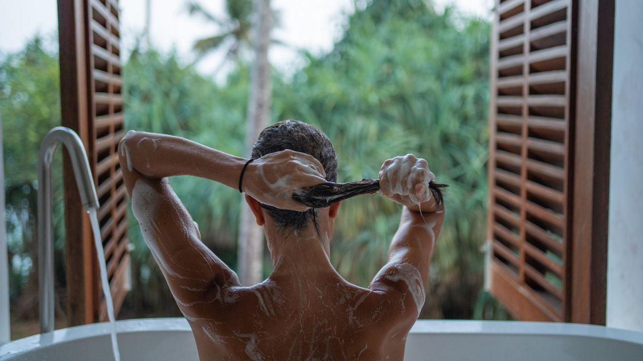 Woman in a bath applying shampoo while looking out at greenery 