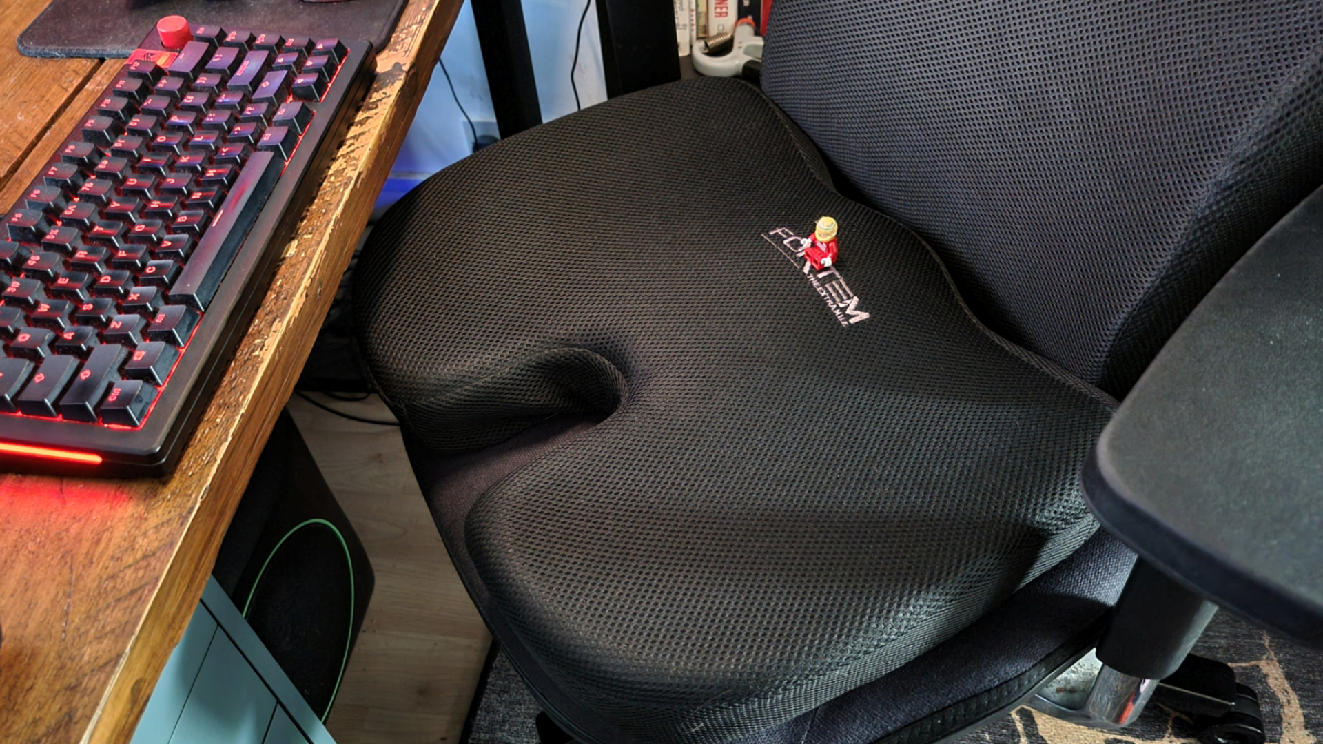 An extra cushion on top of a gaming chair, in front of a gaming keyboard, with a Lego minifig sitting on top