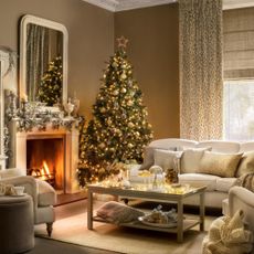 Decorated Christmas tree in a country style brown living room, white sofa, cushions, fireplace, lit fire, decorated mantelpiece, large mirror, coffee table. Pub Orig