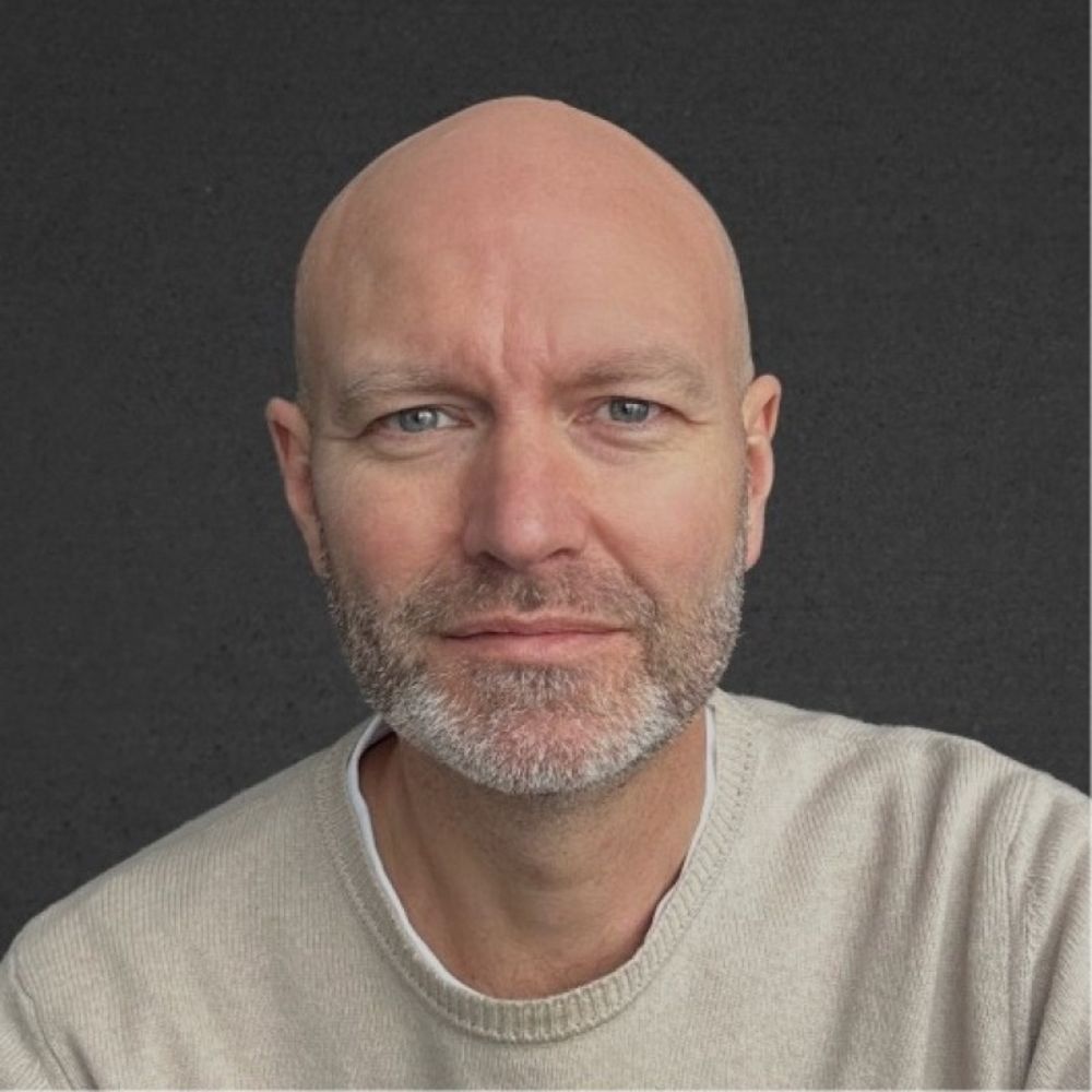 A close-up portrait of a bald man with light stubble, wearing a neutral jumper against a dark background, with soft lighting and a calm, direct expression.