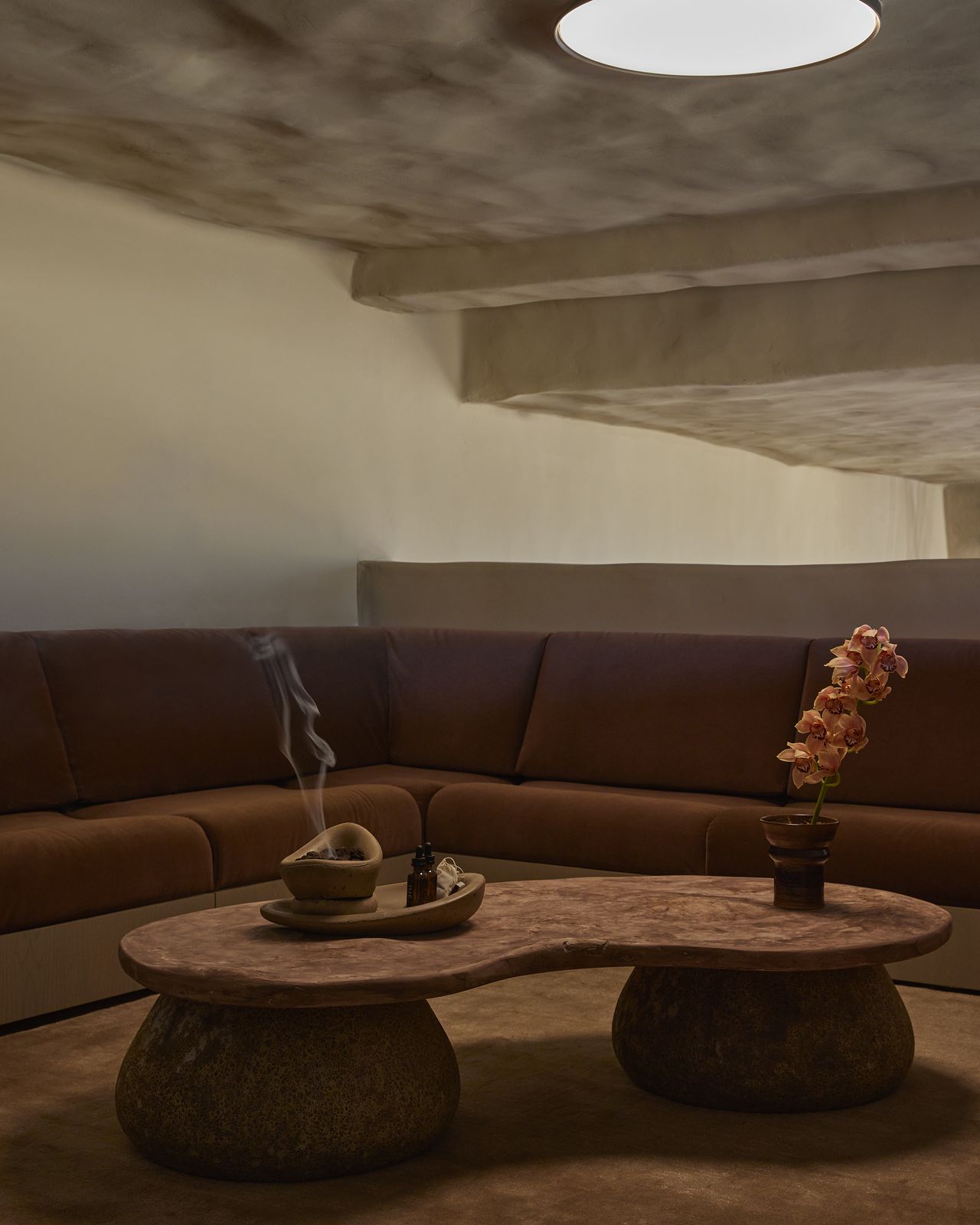 Incense is released into a dimly lit sculptural room filled with brown furniture and lit up by a porthole ceiling window.