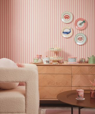 A sitting area with pink striped wallpaper, four plates on the wall, a wooden sideboard with cakes and teacups on it, a cream boucle arm chair and a brown circular table