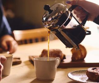 A french press pouring into a cup of coffee on a table with pastires