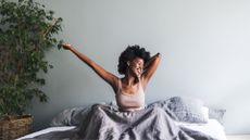 Woman stretching and waking up, smiling in sunlit room with plant next to bed after learning how not to feel tired in the morning