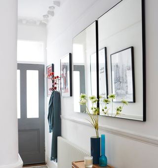 White hallway with grey door with a mirror on teh white wall above a blue vase with white flowers