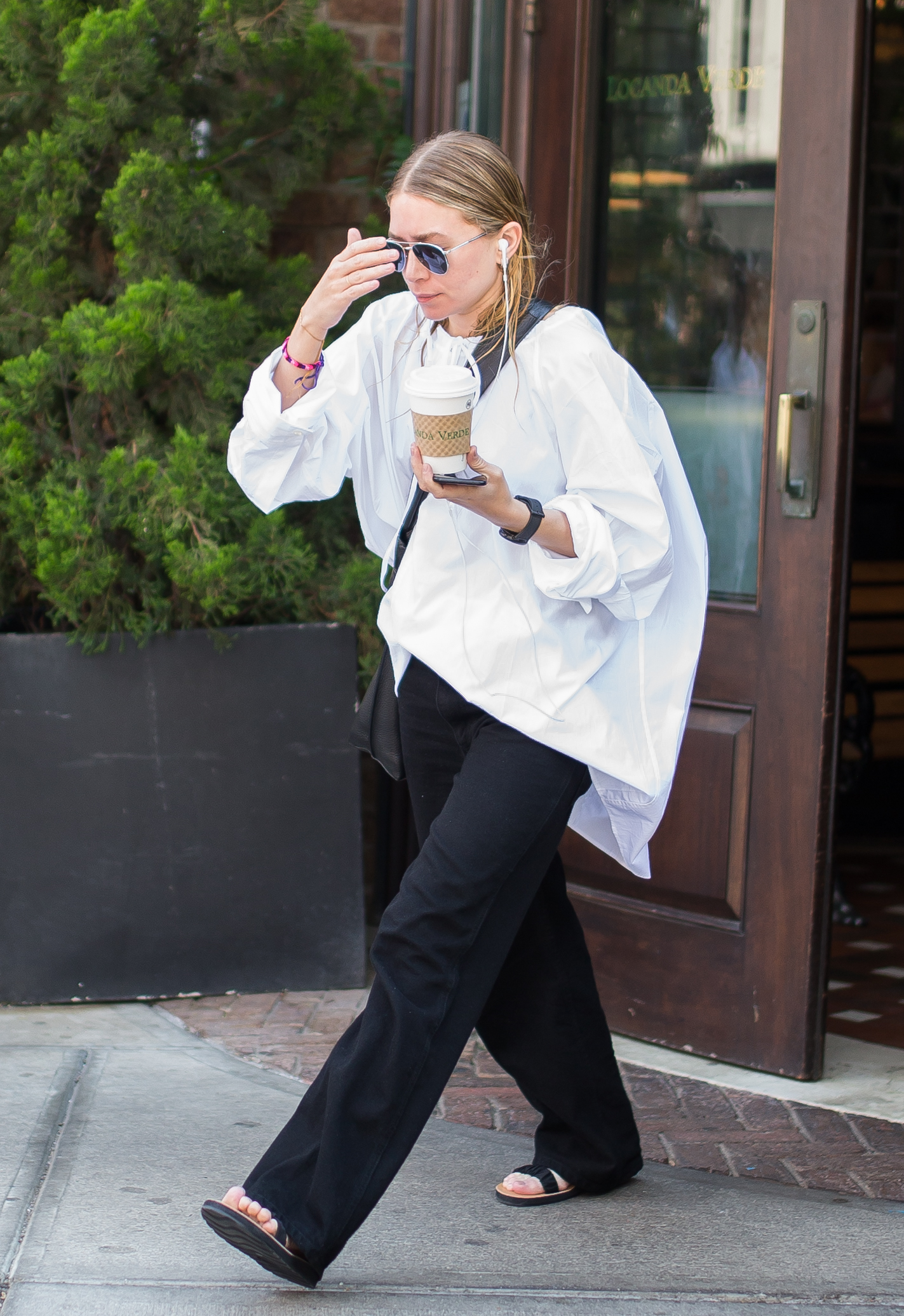 Ashley Olsen, vestindo uma blusa branca de tamanho grande, calça preta e sandálias planas pretas.