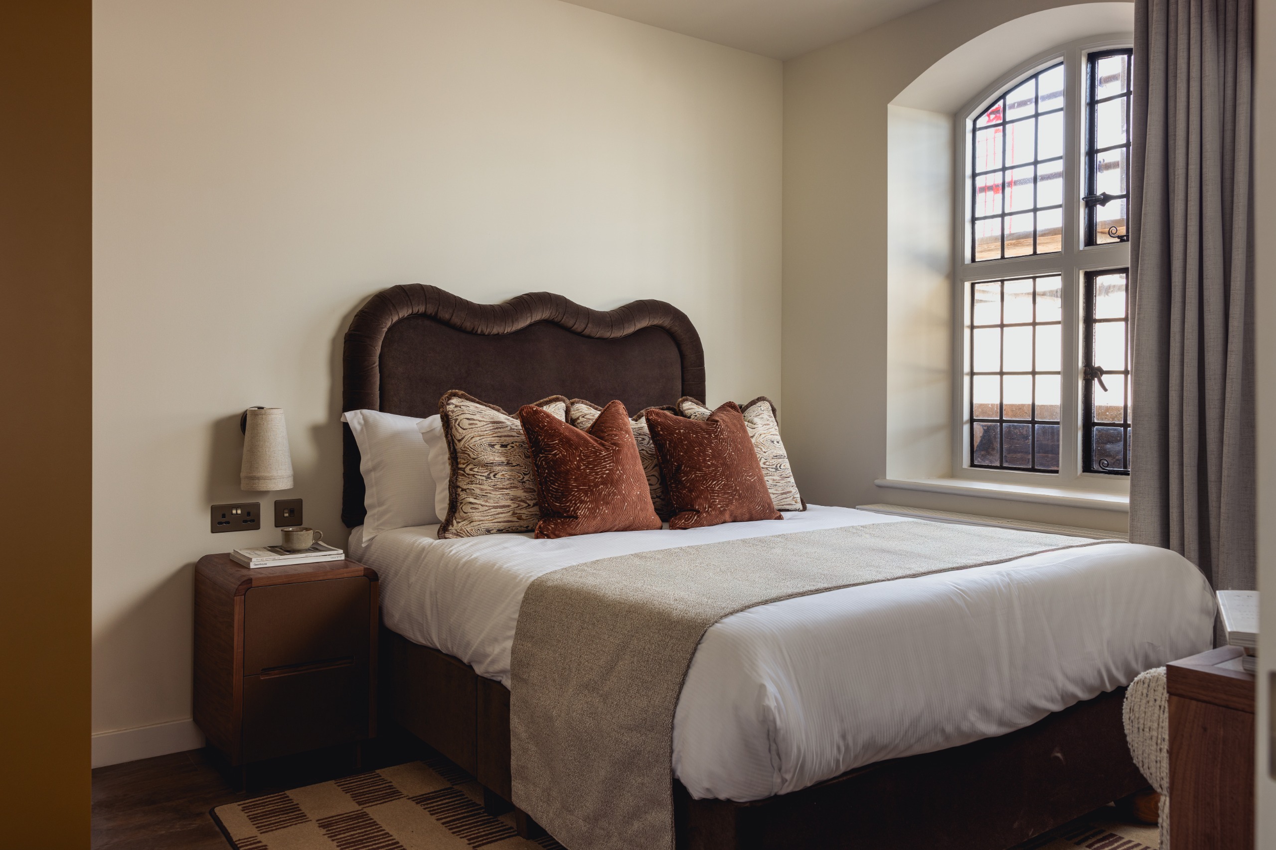 Warm-toned hotel bedroom with wavy, brown velvet headboard, earth red and patterned beige cushions, a beige runner, and checkered neutral rug under the bed and wooden bedside table.