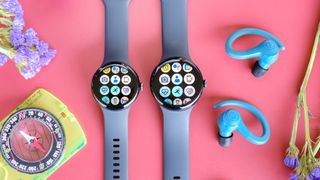 The Google Pixel Watch 3 in 41mm and 45mm sitting beside each other on a red table next to blue earbuds, purple flowers and a yellow compass