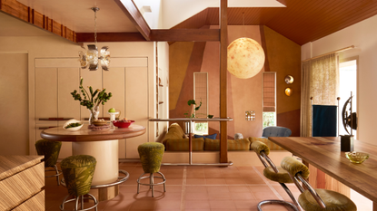 A modern open plan living space with wood and metal accents, a round built-in table with three green stools around it and a large moon-like pendant light about the conversation pit-style seating area at the back