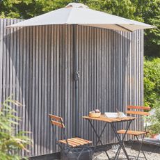 Sweeek half parasol against a panelled wall in a garden next to a wooden bistro set