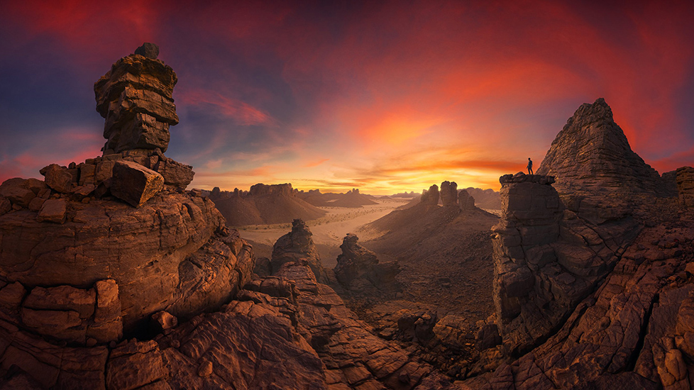A vast desert landscape at sunset, featuring unique rock formations silhouetted against a vibrant sky with a lone figure on a peak