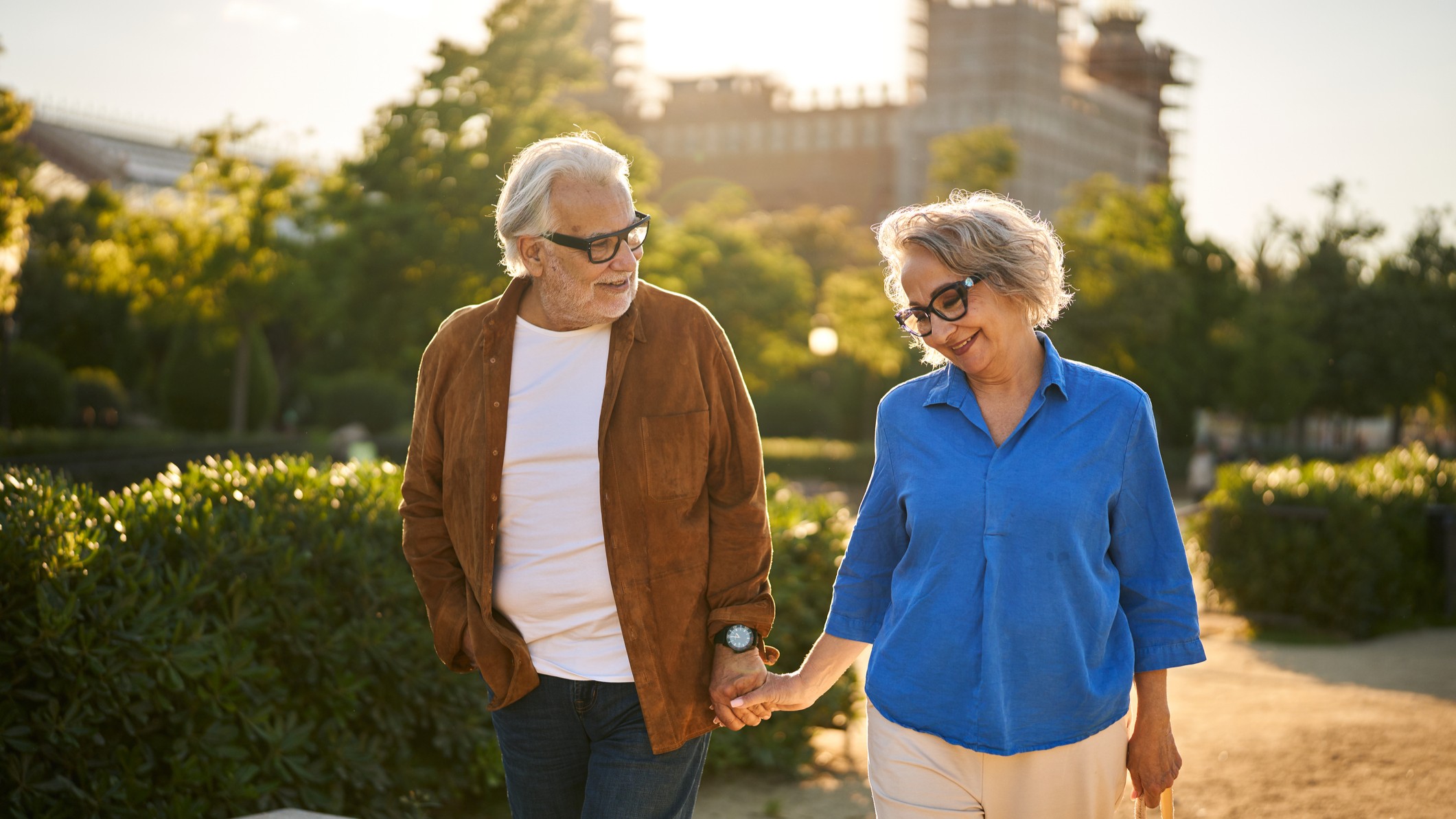An older heterosexual couple walks through a park at golden hour.