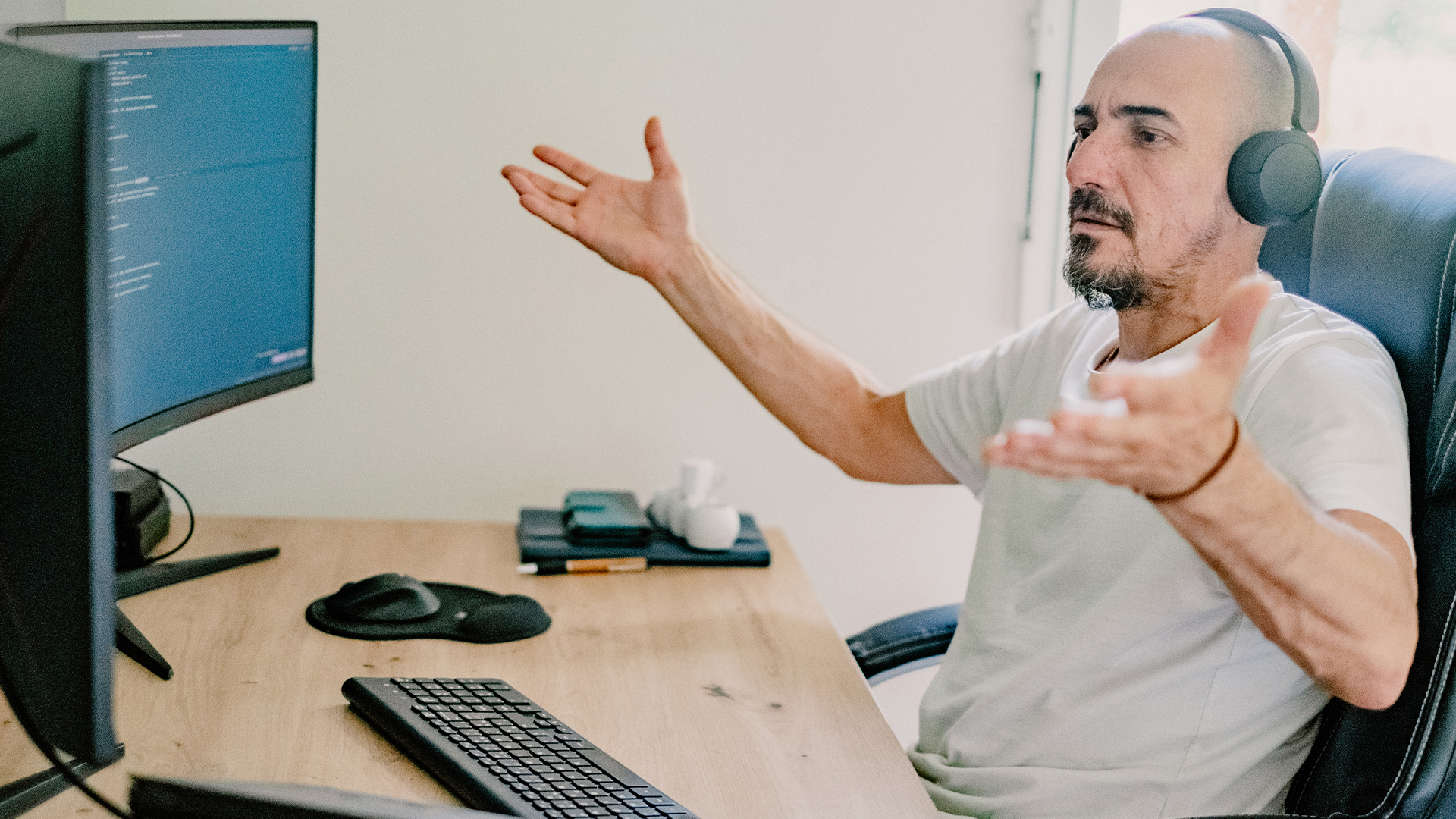 Frustrated male remote worker sitting at desk with headphones on, holding hands up in confusion. 
