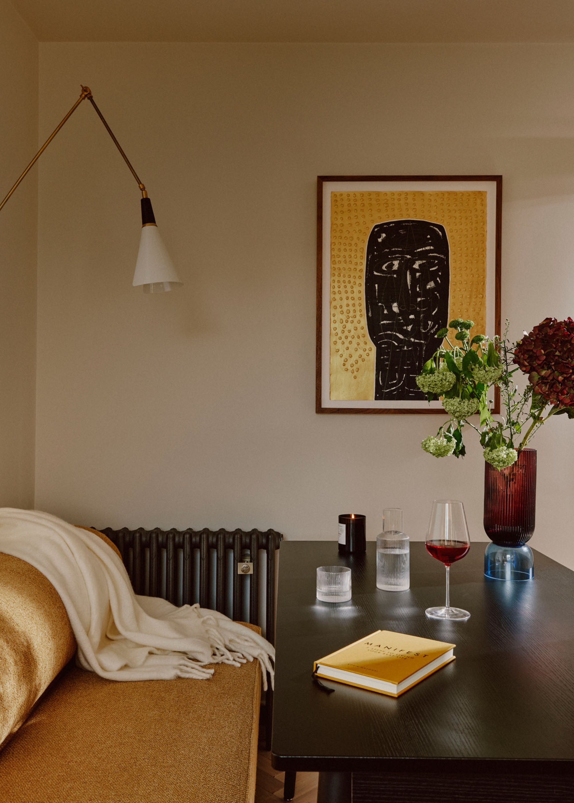 A dining table with a glass of vine, a book and a tall vase with colorful flowers