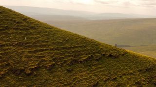 A small lone figure in a backpack hikes up a long, steep hill, attempting the Fan Dance.