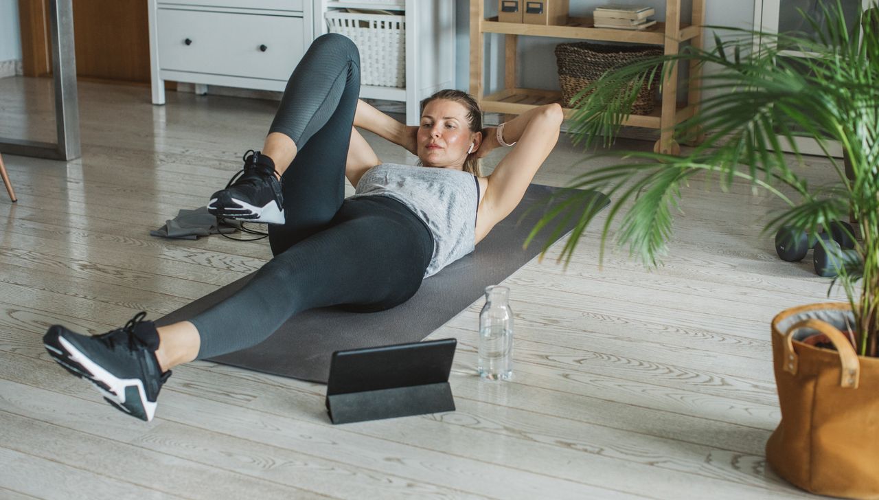 Woman exercising at home. She is lying on her back on an exercise, with her head and shoulders raised off the floor and her hands behind her head. Her legs are lifted and one knee bent and brought towards her chest.