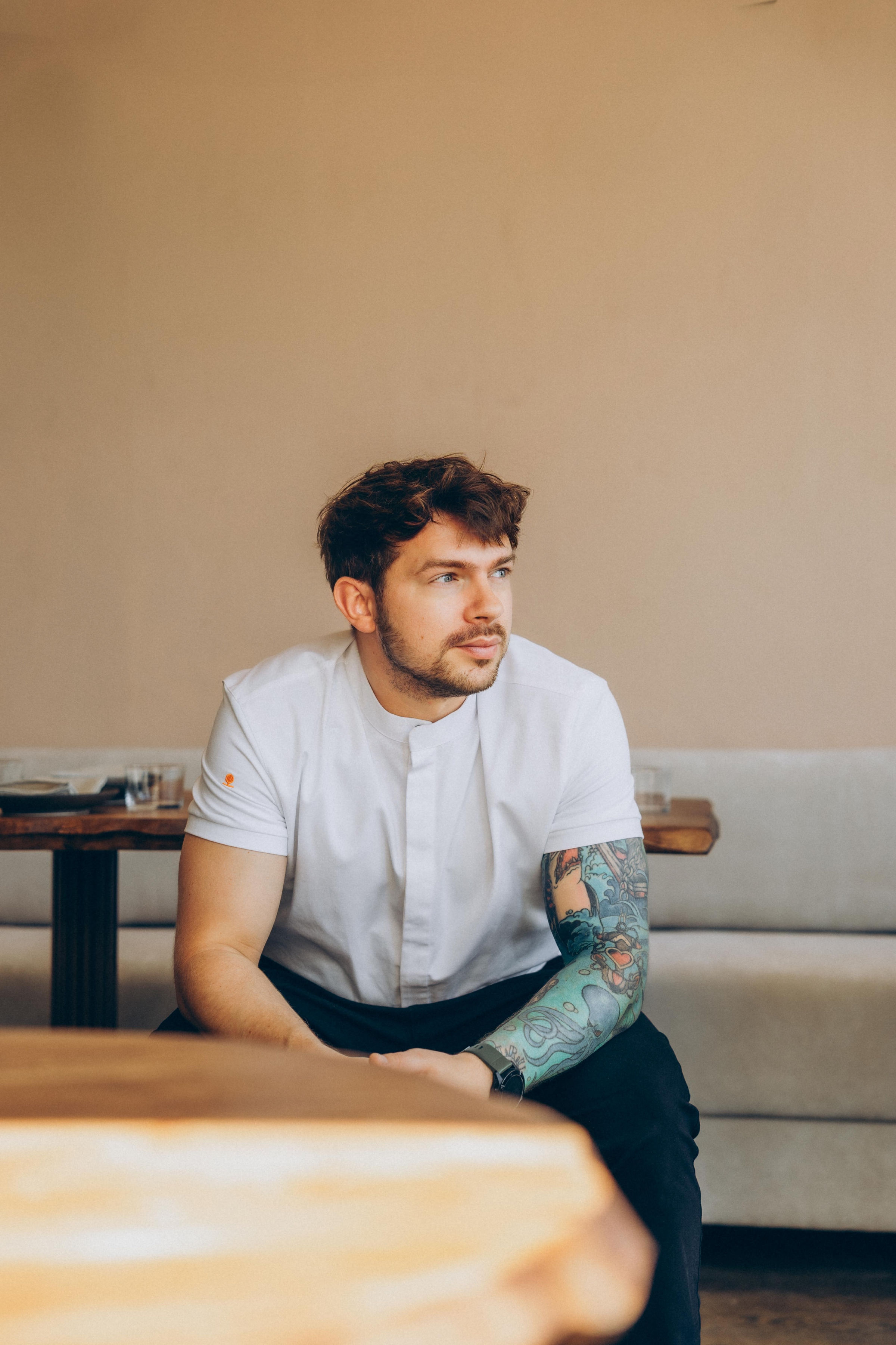 A young male chef dressed in cooking attire sits in a cream-tinted restaurant while staring to his left.