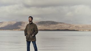 Man wearing wax jacket stares into distance against ocean and mountain backdrop