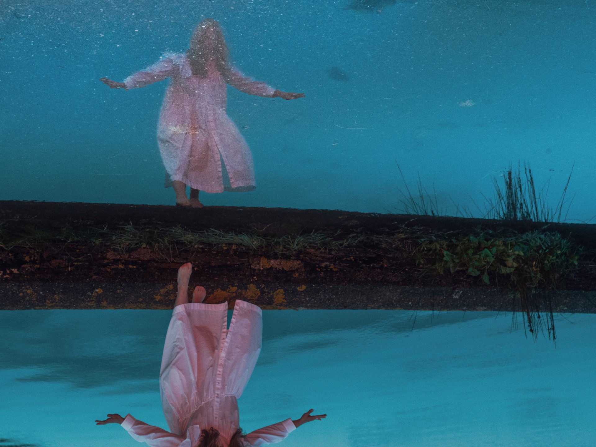 A surreal composition in deep blue tones featuring a figure in a light-colored dress reflected upside down across a dark horizontal divider.