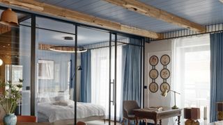 Image of an open-concept living room and bedroom separated by a glass wall with sliding doors. The ceiling is wood-panelled and painted blue.