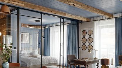 Image of an open-concept living room and bedroom separated by a glass wall with sliding doors. The ceiling is wood-panelled and painted blue.