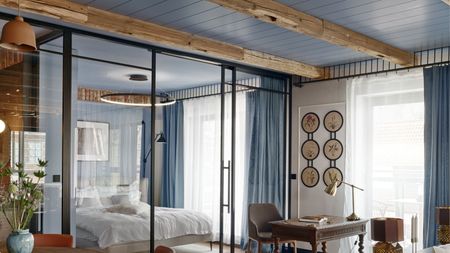 Image of an open-concept living room and bedroom separated by a glass wall with sliding doors. The ceiling is wood-panelled and painted blue.