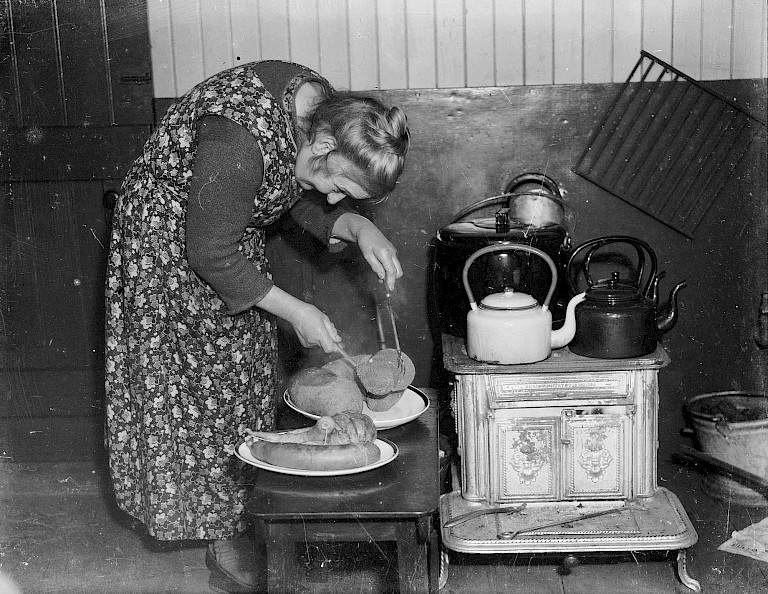 A woman in a floral apron leans over a plate of sheep's puddings beside an old cast-iron kitchen stove.