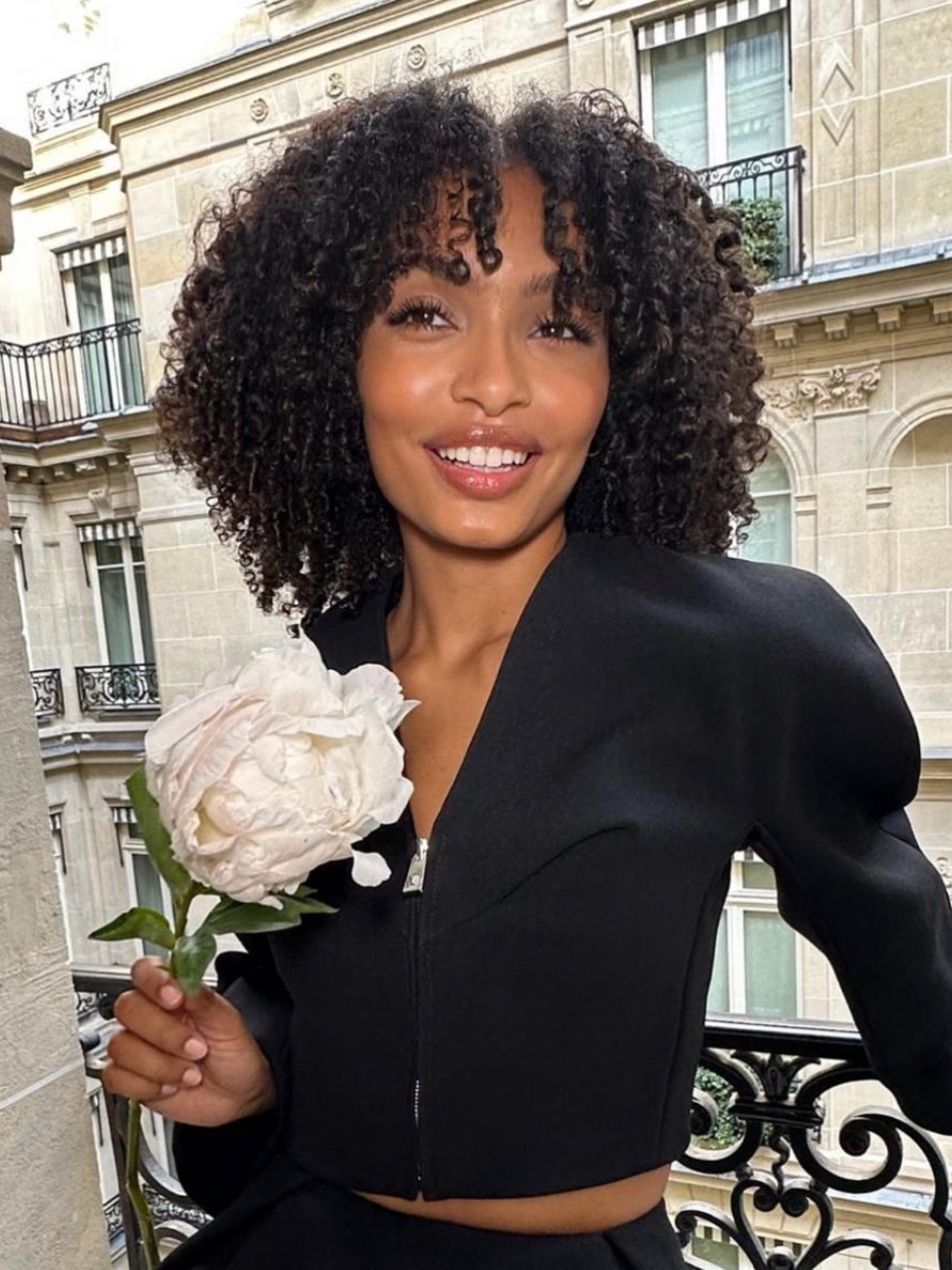 A woman with a curly fringed haircut standing on a balcony holding a flower.