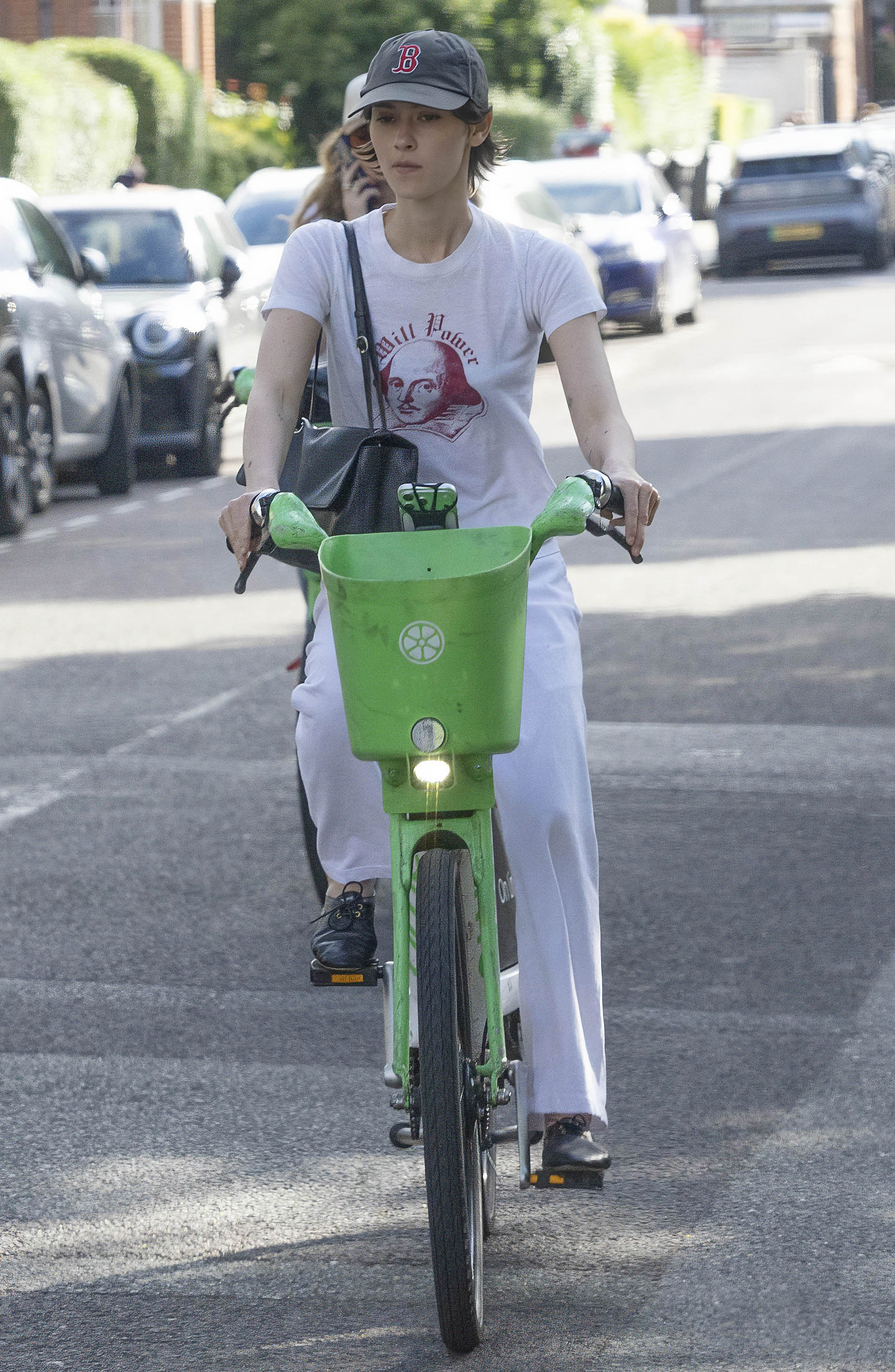 Gracie Abrams cycling through London on a bike wearing a Shakespeare T-shirt, a Boston Red Sox hat, white pants, and black Celine jazz shoes.