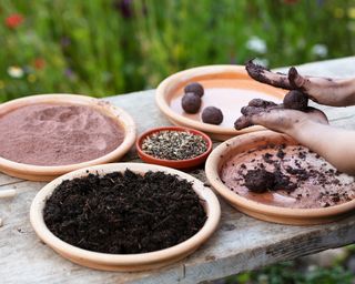 Woman is making seed bombs on a wooden table, using compost and clay powder, with a flower field in the background